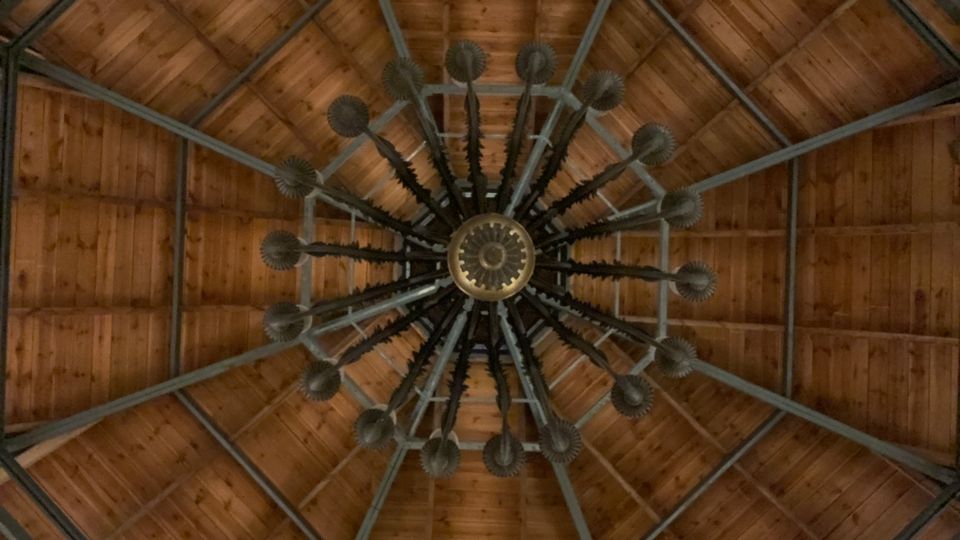 Blick nach oben in eine achteckige Holzdachkonstruktion mit sichtbaren Metallstreben. In der Mitte hängt ein großer, sternförmiger Kronleuchter mit vielen radial angeordneten Armen.
