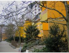 Mehrstöckige Wohngebäude mit gelben Fassaden entlang eines Fußwegs, der von kahlen Bäumen und Büschen gesäumt ist. Im Vordergrund führen Treppen mit Metallgeländern zu den Eingängen, während im Hintergrund weitere Gebäude und Grünflächen sichtbar sind.