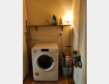 Kleiner Waschraum mit einer weißen Waschmaschine auf einem Holzboden. An der Wand darüber befindet sich ein Regal mit Reinigungsmitteln, daneben eine eingeschaltete Wandlampe; rechts steht eine Plastikbox mit weiteren Haushaltsgegenständen, und eine Seilzugmechanik ist an der Wand montiert.