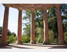 Offener Pavillon mit runden, ziegelroten Säulen und steinernen Basen. Im Hintergrund sind grüne Bäume und ein blauer Himmel zu sehen.