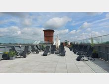 Dachterrasse mit gepflastertem Boden, schwarzen Sonnenliegen und Glasgeländern auf beiden Seiten; im Hintergrund ein roter Backsteinturm und blauer Himmel mit Wolken.