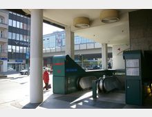 Eingangsbereich zur U-Bahn-Station "Schloßstraße" mit grünen Rolltreppen und Hinweisschildern, umgeben von Straßenszene mit parkenden Autos, Gehweg und modernen Gebäudefassaden im Hintergrund.