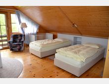 Schlafzimmer mit holzverkleideter Dachschräge, zwei Einzelbetten, einem Sessel, einem Kinderbett und einem kleinen Balkon mit Fenstertüren. Der Raum hat helle Holzböden und Tageslicht dringt durch die Fenster mit gemusterten Vorhängen.