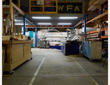 Werkstatt mit Holzverarbeitungsmaschinen und Lagerregalen. Im Vordergrund stehen Arbeitstische und Maschinen, während im Hintergrund Regale mit Materialien und Werkzeugen zu sehen sind.