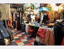 Ein vollgestopfter Kostüm- und Requisitenraum mit Regalen, die mit Perücken, Hüten und Accessoires gefüllt sind, sowie Tischen mit Schminkutensilien und Stoffen. Der Raum hat einen rot-schwarz karierten Boden und wirkt eng durch die große Menge an aufgehängten und gestapelten Gegenständen.