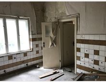 Verlassener Raum mit beschädigten weißen Wandfliesen, teilweise mit braunen Verzierungen. Ein einfaches Fenster mit Blick nach draußen und eine beschädigte Tür prägen die Szene, der Boden ist mit Schutt bedeckt.