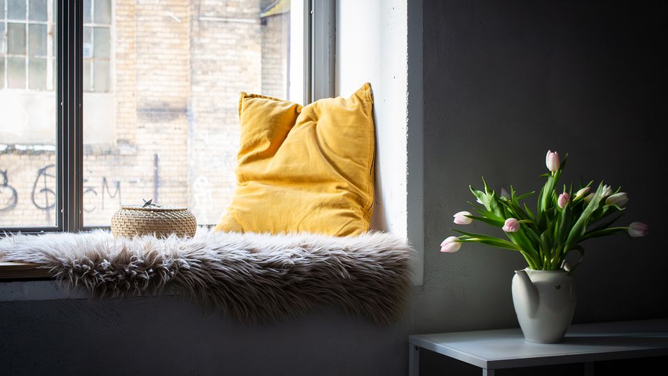 Gemütliche Fensternische in einem Innenraum mit großem Fenster, Fellauflage und gelbem Kissen; auf der Fensterbank steht ein geflochtener Korb. Daneben ein kleiner weißer Tisch mit Vase voller rosa Tulpen vor einer hellen Wand, draußen sind Backsteinfassaden zu sehen.