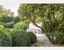 Ein gepflegter Garten mit dichten Sträuchern, einer kleinen Kiesfläche und einer modernen weißen Sitzbank mit lila Polsterung, daneben ein Baum mit breitem Stamm und überhängenden Zweigen. Im Hintergrund sind hohe Hecken zu sehen.