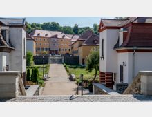 Hof einer historischen Schlossanlage mit gelb gestrichenen Gebäuden und roten Dächern; in der Mitte führt ein gepflasterter Weg vorbei an Grünflächen zu einer Treppe, die zum Hauptgebäude mit verziertem Eingangsportal führt. Links und rechts sind flankierende Gebäude mit weißen Fassaden und roten Dächern zu sehen.