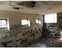 Innenraum eines verlassenen Gebäudes mit verwitterten Ziegelwänden, teils abblätterndem Putz und kleinen rechteckigen Fenstern, durch die Tageslicht einfällt. Der Boden ist bedeckt mit Laub und Schutt, und Spuren von Feuchtigkeit und Verfall sind an Wänden und Decke erkennbar.