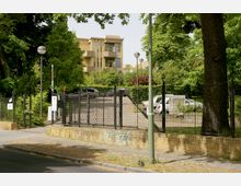 Gepflasterter Gehweg vor einem Metallzaun mit Maschendraht, der eine begrünte Parkfläche mit geparkten Autos umgibt. Im Hintergrund ist ein mehrstöckiges gelbes Gebäude mit Balkonen und großen Fenstern zu sehen, umgeben von Bäumen und weiteren Pflanzen.