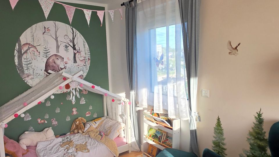 Kinderzimmer mit Holzfußboden, Hausbett und grüner Akzentwand mit großem Waldtier‑Wandbild sowie Wimpelgirlanden und einer weißen Deckenlampe. Ein großes Fenster mit halbtransparenten Vorhängen, ein kleines Regal und ein dunkelgrüner Sessel ergänzen die Einrichtung.
