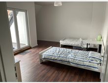 Ein schlicht eingerichtetes Schlafzimmer mit zwei Einzelbetten, einem kleinen Schreibtisch mit Stuhl und weiß gestrichenen Wänden. Der Raum hat einen dunklen Holzfußboden und Zugang zu einem Balkon durch eine große Fenstertür.