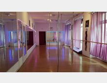 Ein Tanzstudio mit Holzfußboden, mehreren vertikalen Stangen und großen Wandspiegeln. Die Fenster sind mit lila Vorhängen bedeckt, und bunte LED-Leuchten beleuchten die Wände.