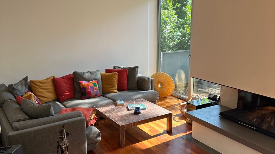 Helles, modernes Wohnzimmer mit großem grauen Ecksofa und bunten Kissen um einen niedrigen Couchtisch auf Holzfußboden. Rechts befindet sich ein moderner Einbaukamin mit Glasfront, daneben ein bodentiefes Fenster mit Blick ins Grüne.
