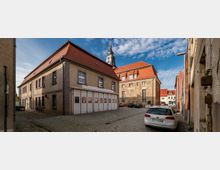 Historischer Innenhof mit Kopfsteinpflaster, umgeben von Gebäuden mit roten Ziegeldächern, darunter ein größeres Gebäude mit einer Kirche und Glockenturm im Hintergrund. Geparkte Autos und klare, sonnige Wetterbedingungen prägen die Szene.