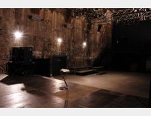 Industriehalle mit einer Bühne und rauen, unverputzten Wänden, die mit Graffiti bedeckt sind. Im Vordergrund ein einzelner Hocker, im Hintergrund Lautsprecher, Podeste und eine Metalltreppe, ergänzt durch eine Deckenbeleuchtung mit Scheinwerfern an einem Gerüst.