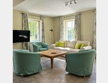 Helles Wohnzimmer mit beigefarbenen Wänden, zwei Fenstern mit gemusterten Vorhängen, einem weißen Sofa mit grünen Kissen, zwei grünen Sesseln und einem Holztisch in der Mitte. An der linken Wand hängt ein Flachbildfernseher, daneben steht eine Stehlampe.