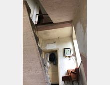 Treppenhaus in einem alten Gebäude mit sichtbaren Betonwänden und abblätterndem Putz. Im Vordergrund stehen ein orangefarbener Sessel und ein gerahmtes Bild an der Wand, während durch eine offene Tür im Hintergrund Kleidung und ein Rucksack sichtbar sind.