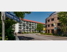 Straßenszene mit einem geschwungenen Holzgebäude und einem mehrstöckigen Wohnhaus mit Balkonen und rotem Dach im Hintergrund. Ein Straßenschild mit der Aufschrift "Geschwister-Scholl-Straße" ist links im Vordergrund sichtbar, umgeben von Bäumen und Grünflächen.