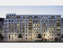 Großes, mehrstöckiges Wohngebäude mit weißer Fassade, zahlreichen Balkonen und großen Fenstern, die größtenteils beleuchtet sind. Vor dem Gebäude stehen Straßenlaternen und einzelne Bäume entlang des Gehwegs.