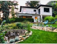 Zweistöckiges Haus mit heller Fassade, Flachdach und Balkon, umgeben von einem großen Garten mit Blumenbeeten, Sträuchern und einem Steingarten. Im Vordergrund befindet sich eine Sitzgruppe mit Holzstühlen und ein kleines überdachtes Gartenareal.