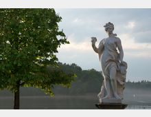 Steinfigur einer weiblichen Gestalt in fließender Kleidung neben einem großen, grünen Baum am Ufer eines ruhigen Sees mit bewaldeter Hintergrundlandschaft unter bewölktem Himmel.
