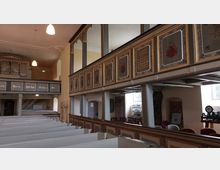 Innenraum einer historischen Kirche mit weiß gestrichenen Holzbänken, verzierten Emporen mit Gemälden und Schriftzügen sowie einer kleinen Orgel im Hintergrund. Die Wände sind in gedeckten Farben gehalten, und durch Fenster fällt Tageslicht ein.