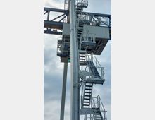 Metallkonstruktion einer großen Industriemaschine mit mehreren Etagen von Treppen und Plattformen, die durch Geländer gesichert sind. Im Hintergrund ist ein wolkiger Himmel zu sehen.