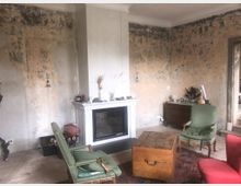 Wohnzimmer mit rustikalem Charakter, gekennzeichnet durch verblasste, teilweise abgeblätterte Wände. Im Vordergrund stehen Sessel, ein Holz-Couchtisch und ein roter Teppich, während ein moderner Kamin und eine schlichte weiße Kaminkonsole die Rückwand dominieren.