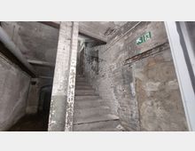 Treppenhaus mit abgenutzten Betonstufen in einem alten, verlassen wirkenden Kellerbereich mit unverputzten Ziegelwänden. Im Hintergrund befindet sich ein Notausgangsschild an der Wand, und oben führen die Stufen zu einer Öffnung.