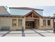 Eingang eines modernen Gebäudes mit gelblicher Backsteinfassade, Holzrahmenfenstern und einem kleinen Vordach aus Holz und Ziegel. Im Vordergrund steht ein Fahrrad vor einem Fahrradständer auf dem gepflasterten Vorplatz.