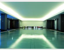 Schwimmbad in einem modern gestalteten Innenraum mit glatter Wasseroberfläche, niedriger Decke mit indirekter Beleuchtung und rechteckigen Glasfenstern am Ende des Raumes.