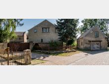 Straßenszene mit einem alten Wohnhaus mit verputzter Fassade und Satteldach, einem angrenzenden kleineren Gebäude mit verfallenem Tor und Graffiti sowie einer gepflasterten Straße im Vordergrund. Der Bereich ist von Bäumen und niedrigem Metallzaun umgeben.