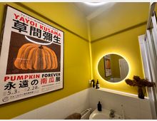 Kleines Badezimmer mit gelb gestrichenen Wänden, einem beleuchteten runden Spiegel und einem Waschbecken mit weißen Fliesen. An der Wand hängt ein Plakat mit der Aufschrift "Pumpkin Forever" und einer Abbildung eines gepunkteten Kürbisses, daneben stehen Dekorationsobjekte und ein getrockneter Blumenstrauß.
