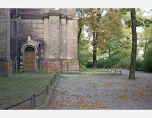 Außenansicht einer historischen Backsteinmauer mit schmalem Portal, verziert mit einem steinernen Bogen. Rechts daneben liegt ein parkähnlicher Bereich mit Bäumen, Pflastersteinen und Laub auf dem Boden.