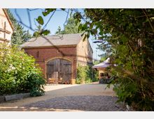 Ansicht eines ländlichen Hofs mit einem historischen Backsteingebäude und großen Holztoren in der Mitte. Im Vordergrund pflasterter Weg, Blumenbüsche und ein kleiner Sitzbereich, rechts ein Verkaufsstand mit Zelt.