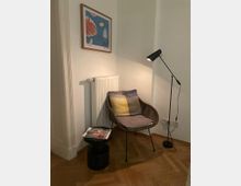 Eine gemütliche Ecke in einem Raum mit einem geflochtenen Stuhl, darauf ein Kissen und eine Decke, daneben ein kleiner runder Beistelltisch mit einer Zeitschrift und eine schwarze Stehlampe. An der Wand hängt ein gerahmtes Bild, und unterhalb ist ein Heizkörper montiert. Der Boden besteht aus Parkett.