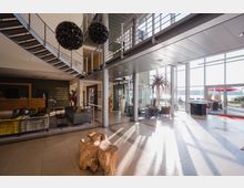 Heller, moderner Lobbybereich eines Gebäudes mit hohen Glasfassaden, die Tageslicht einlassen. Zu den Hauptmerkmalen gehören eine offene Treppe mit Metallgeländer, Sitzbereiche mit Sofas und Sesseln, dekorative Beleuchtung und eine hölzerne Rezeption.