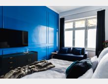 Schlafzimmer mit einer intensiv blauen Akzentwand, einem Flachbildfernseher, einer schwarzen Kommode und einem Bett mit weißer Bettwäsche und einer Kunstfelldecke. Im Raum befindet sich auch ein Sofa vor einem großen Fenster mit weißen Vorhängen und dunklen Gardinen.