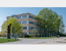 Ein beige Mehrzweckgebäude mit mehreren Etagen und vielen Fenstern, umgeben von Bäumen und einem Zaun. Im Vordergrund führt ein gepflasterter Weg mit zwei Personen daran entlang, während ein Baumstumpf links im Bild sichtbar ist.