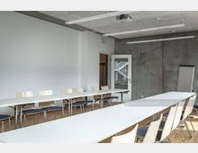 Konferenzraum mit langen weißen Tischen und hellen Holzstühlen auf einem Boden aus Holzlaminat. An der Wand befinden sich eine Glastür, ein Flipchart und eine Betonwand kombiniert mit weißen Elementen.