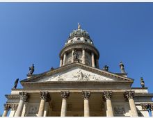 Frontale Ansicht eines historischen Gebäudes mit säulenverzierter Fassade und figürlichem Relief im Dreiecksgiebel. Eine Kuppel mit einer vergoldeten Statue und Statuen auf dem Dach dominieren das Bild, der Himmel ist klar und blau.