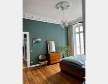 Schlafzimmer mit dunkelgrüner Wand, verziert mit weißem Stuck an der Decke. Im Raum befinden sich ein Holzbett, eine Kommode mit einem goldgerahmten Spiegel, ein Pflanzenkübel, ein Holzstuhl und ein Kronleuchter mit Glaskugeln. Der Boden besteht aus poliertem Holz, und ein großes Fenster mit cremefarbenen Vorhängen sorgt für Tageslicht.