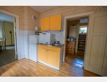 Kleine, ältere Küche mit gelben Hängeschränken, weißen Fliesen an der Wand, Kühlschrank, Spüle und einer kompakten Spülmaschine auf der Arbeitsfläche. Gemusterte Tapete im Retro-Stil, Laminatboden und offene Türdurchgänge zu angrenzenden Räumen.