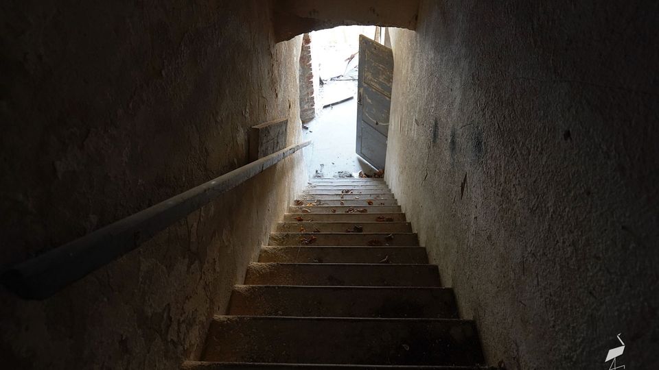 Blick von oben in ein schmales, abgenutztes Treppenhaus mit Betonstufen und rauen Putzwänden, links mit einfachem Handlauf. Unten führt eine halb geöffnete Tür durch einen niedrigen Rundbogen ins helle Außenlicht.