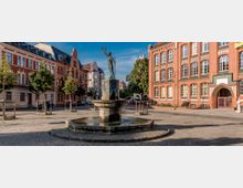 Ein gepflasterter Platz mit einem zentralen Brunnen, auf dem eine Statue steht, die eine erhobene Hand mit einem Kranz zeigt. Im Hintergrund befinden sich Backsteingebäude mit verzierter Architektur sowie Wohnhäuser, die von Bäumen gesäumt sind.