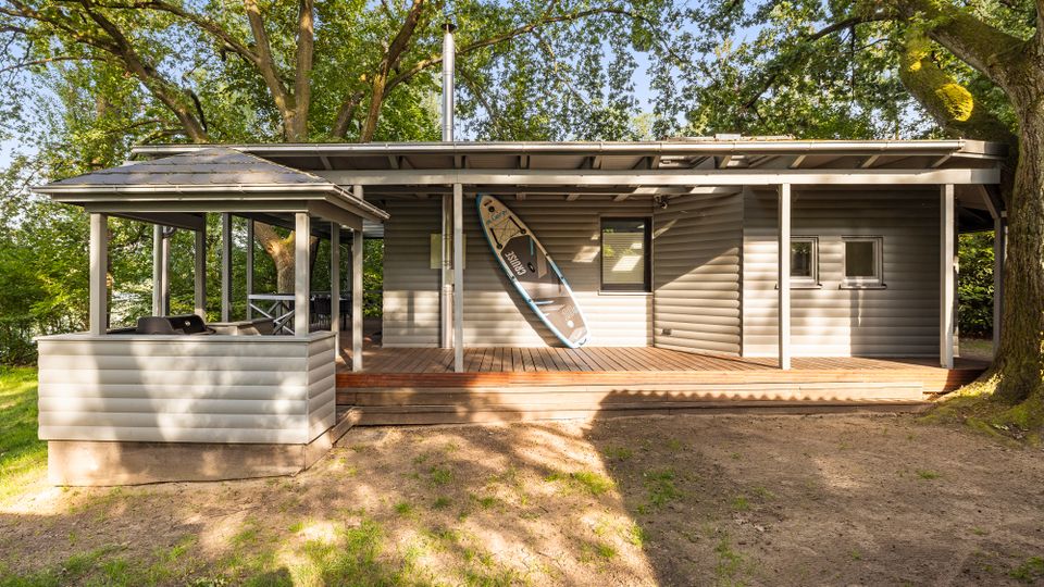 Modernes, eingeschossiges Holzhaus mit breiter, überdachter Veranda und Holzterrasse, umgeben von großen Bäumen. Seitlich befindet sich ein offener Pavillonbereich, und an der Hauswand lehnt ein Stand-up-Paddleboard.