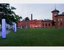 Backstein-Bauensemble mit rotem Ziegelmauerwerk, bestehend aus mehreren Gebäuden, darunter ein Turm und ein Schornstein, beleuchtet in warmem Licht. Im Vordergrund eine Rasenfläche mit wenigen Leuchtsäulen, im Hintergrund versammeln sich Menschen vor den Gebäuden.