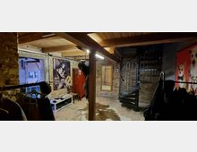 Raum mit industriellem Design, gekennzeichnet durch freiliegende Ziegelwände und eine Holzdecke. Im Vordergrund befinden sich Kleiderständer, eine Wendeltreppe aus Metall führt in eine obere Etage, und im Hintergrund sind Poster, ein großer Spiegel und ein roter Schrank zu sehen.